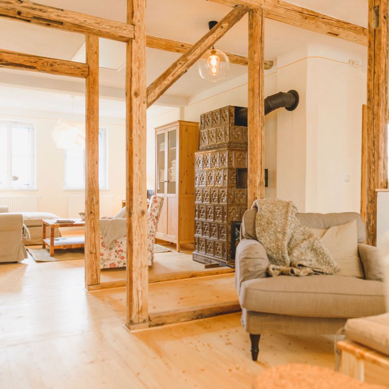 Helles, offenes Wohnzimmer mit vielen Holzbalken, Sessel im Vordergrund. Im Hintergrund ein Kachelofen, Schrank und Sofa mit Tisch.