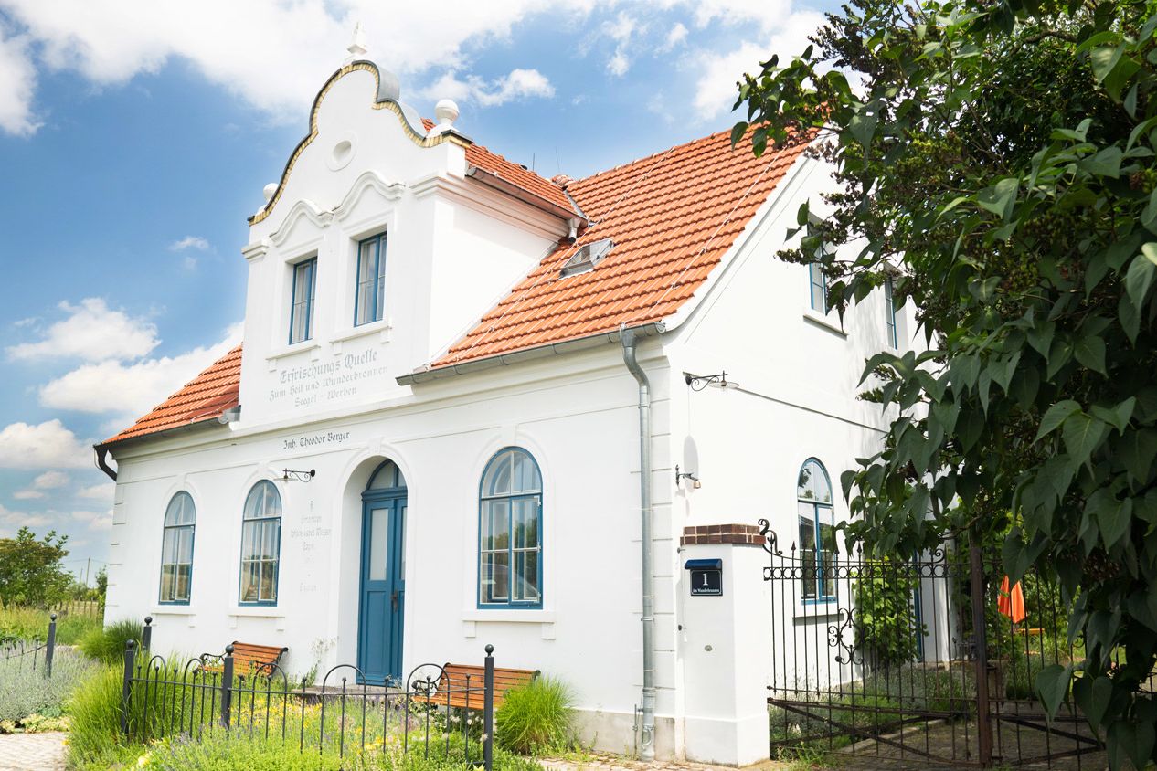 Totale Ferienhaus: weißes Haus mit rotem Ziegeldach und blauer Tür, davor zwei Bänke im Grünen.